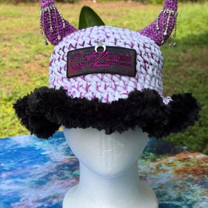 May include: A white and purple crocheted bucket hat featuring two purple horns adorned with silver rhinestone fringe. The hat has a black fuzzy brim and a rectangular patch on the front with the text CLOZEE and a small silver crescent moon charm.