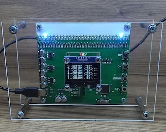 Puede incluir: Una placa de circuito verde con una pequeña pantalla que muestra texto, montada dentro de un marco de acrílico transparente. La placa tiene varios componentes, incluyendo un puerto USB y botones etiquetados "UP", "OK", "DOWN" y "ESC".