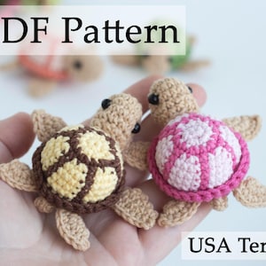 Peut inclure: Deux jouets amigurumi en forme de tortue crochetés. L'un est brun avec des accents jaunes et l'autre est rose et blanc. L'image comprend le texte "PDF Pattern" et "USA Terms".