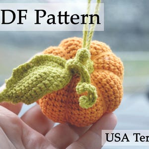 May include: A crocheted pumpkin with a green stem and leaves. The pumpkin is orange and the leaves are green. The text "PDF Pattern" is at the top of the image. The text "USA Terms" is at the bottom of the image.