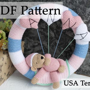 Op de afbeelding: Een gehaakte krans in roze, blauw en wit met een gehaakte teddybeer die eraan hangt. De krans heeft een hartvormig ontwerp en de tekst "PDF Pattern" staat erboven. De tekst "USA Terms" staat onder de krans.