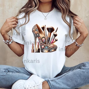 Puede incluir: Camiseta blanca con un gráfico de un zapato de tacón alto dorado con pinceles de maquillaje y lápiz labial en su interior.