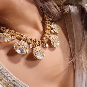 May include: A gold-toned necklace with large, clear, teardrop-shaped pendants. The necklace features a thick chain design with smaller, clear stones. The necklace is worn over a white garment with gold trim.