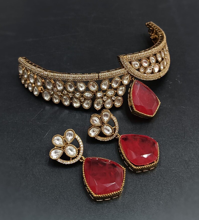 Op de afbeelding: Een gouden chokerketting met een rode edelsteenhanger en bijpassende oorbellen. De ketting en oorbellen zijn versierd met heldere zirkonia-stenen.