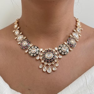 May include: A gold necklace with a green and red enamel design. The necklace features a central pendant with a round shape and a smaller pendant with a teardrop shape. The necklace is adorned with white stones.