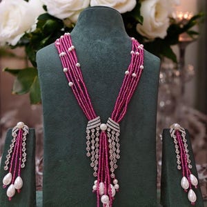 May include: A magenta beaded necklace and matching earrings set displayed on a green velvet bust. The necklace features multiple strands of magenta beads, accented with white pearls and diamond-like embellishments. The earrings have a similar design.