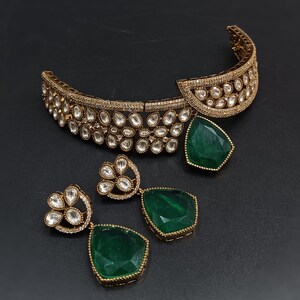 Op de afbeelding: Een gouden choker ketting met een groene edelsteen hanger en bijpassende oorbellen. De ketting is versierd met heldere stenen en de oorbellen hebben een bloemmotief.