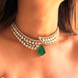May include: A gold choker necklace with a large emerald green gemstone pendant. The necklace is made of small, round, clear stones.