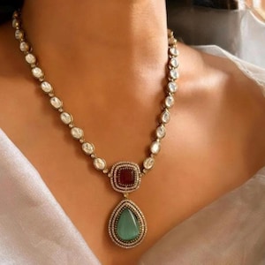 May include: A gold necklace with a teardrop-shaped pendant featuring a green gemstone surrounded by smaller clear stones. The pendant is attached to a square red gemstone surrounded by clear stones. The necklace is made of a chain of clear stones.