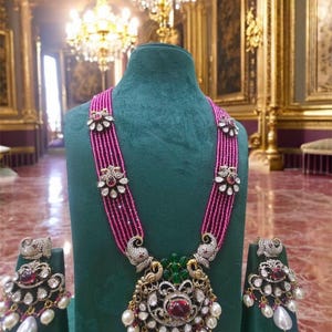 May include: A pink and white beaded necklace with a large pendant featuring a peacock design and red and green gemstones. The necklace is displayed on a green velvet stand with matching earrings.