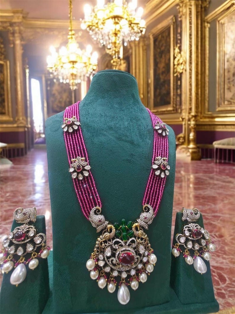 May include: A pink and white beaded necklace with a large pendant featuring a peacock design and red and green gemstones. The necklace is displayed on a green velvet stand with matching earrings.