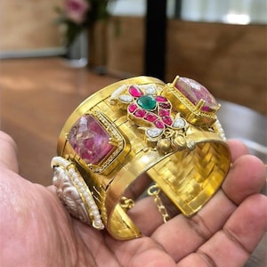 May include: A gold-coloured cuff bracelet with large, rectangular pink gemstones and smaller, colourful stones. The bracelet has intricate detailing and is held in a hand.