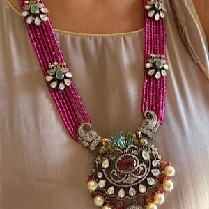 May include: A long, layered necklace with a large, ornate pendant. The necklace is made of pink beads and features a silver pendant with a red gemstone and white pearls. The pendant is decorated with intricate details and is a beautiful example of traditional Indian jewelry.