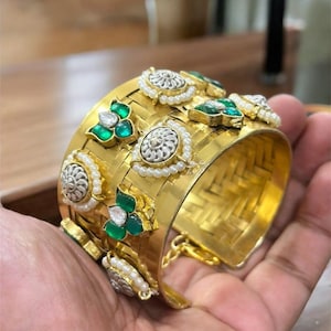 May include: A gold cuff bracelet with detailed ornamentation. The bracelet has a woven pattern and is decorated with silver-coloured circular elements, green gemstones, and tiny pearls. The design is elaborate and striking.