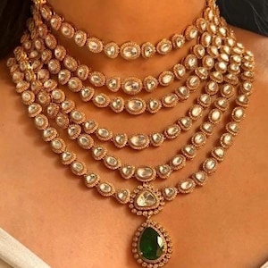 Op de afbeelding: Een gelaagde gouden ketting met een druppelvormige hanger van een groene edelsteen. De ketting is gemaakt van meerdere strengen kleine, ronde, heldere stenen.