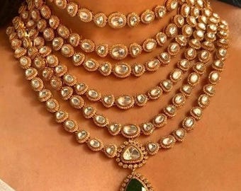Pode incluir: Um colar de ouro em camadas com um pingente de pedra preciosa verde em forma de lágrima. O colar é feito de várias camadas de pequenas pedras redondas transparentes.