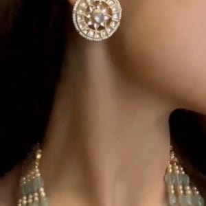May include: A pair of round, gold-toned earrings with a white stone center and a white pearl border. The earrings are worn by a person with dark hair.