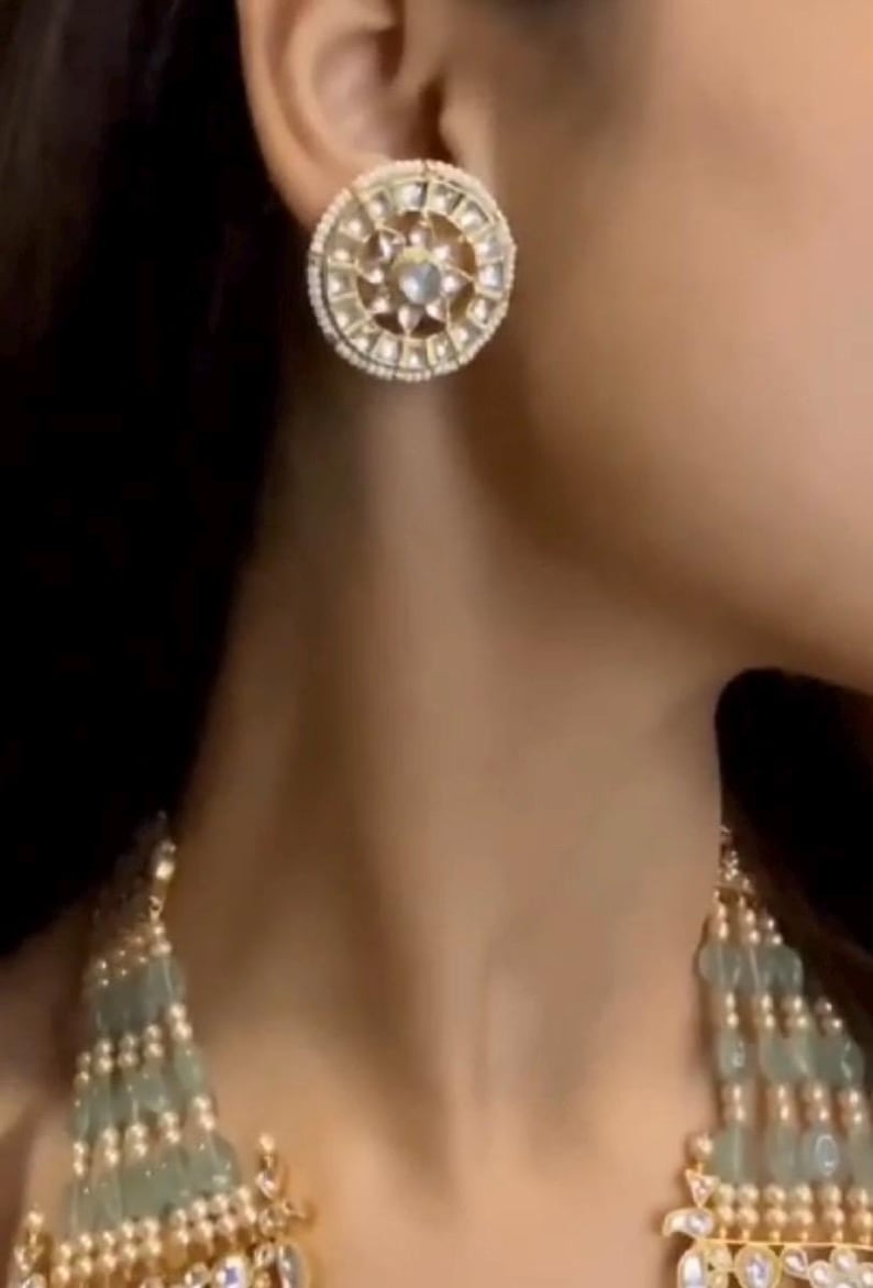 May include: A pair of round, gold-toned earrings with a white stone center and a white pearl border. The earrings are worn by a person with dark hair.