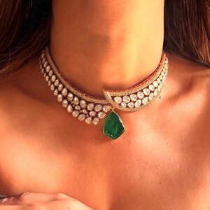 Op de afbeelding: Een gouden choker ketting met een grote smaragdgroene edelsteen hanger. De ketting is versierd met kleine, heldere edelstenen.