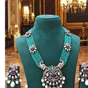 May include: A turquoise and silver necklace with a large pendant featuring a peacock design. The pendant is adorned with white pearls, green stones, and a large red stone. The necklace is displayed on a green velvet mannequin. Matching earrings are also shown.