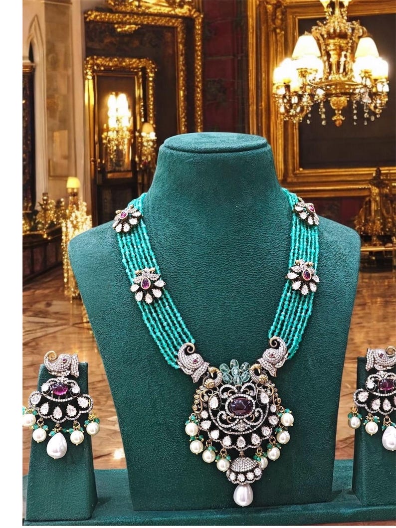 May include: A turquoise and silver necklace with a large pendant featuring a peacock design. The pendant is adorned with white pearls, green stones, and a large red stone. The necklace is displayed on a green velvet mannequin. Matching earrings are also shown.