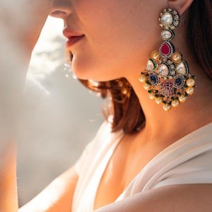 Pode incluir: Um par de brincos grandes e ornamentados com uma base dourada e uma variedade de pedras preciosas coloridas, incluindo vermelho, azul e verde. Os brincos são decorados com pérolas e têm a forma de uma lágrima.