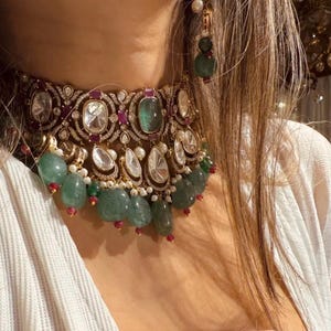 May include: A gold choker necklace with a large emerald green gemstone, surrounded by smaller diamonds and ruby red stones. The necklace is adorned with a fringe of emerald green and white pearls.