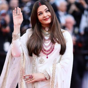 May include: A woman in a white outfit with gold trim, waving to the camera. She wears a red bindi, a choker, and layered necklaces with red accents. Her ring is red and square.