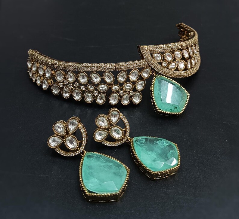 Op de afbeelding: Een gouden choker ketting met een traanvormige groene stenen hanger en bijpassende oorbellen. De ketting is versierd met heldere stenen in een bloemmotief.