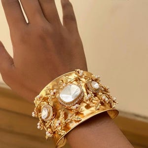 May include: A wide, gold-coloured cuff bracelet with large, clear, faceted stones and smaller, round, white stones. The bracelet has a floral and leaf design with the word "Handmade" in black text.