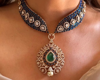 May include: A gold necklace with a large, ornate pendant featuring a green gemstone and blue and white stones. The necklace is made of gold and has a delicate design.