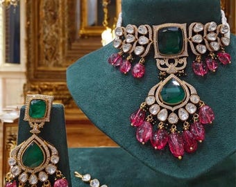 May include: A gold and silver necklace with a large green gemstone pendant and pink beads. The necklace is on a green velvet display stand. There are matching earrings and a maang tikka (forehead ornament) with a green gemstone and pink beads.
