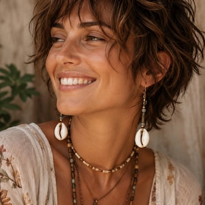 Puede incluir: Una persona con pendientes de concha y collares superpuestos. Los pendientes tienen un colgante de concha blanca. Los collares incluyen hilos de cuentas y un pequeño colgante. Lleva una blusa floral.