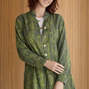 May include: A long-sleeved green tunic with a mottled pattern and wooden buttons. The tunic is worn over a white top and paired with white pants. The garment has a mandarin collar and side slits.