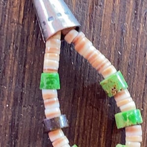 May include: A handcrafted necklace featuring a silver cone pendant and a beaded design. The necklace incorporates small, flat, off-white shell beads interspersed with vibrant green and brown square beads. The beads are strung together, creating a unique and eye-catching accessory.