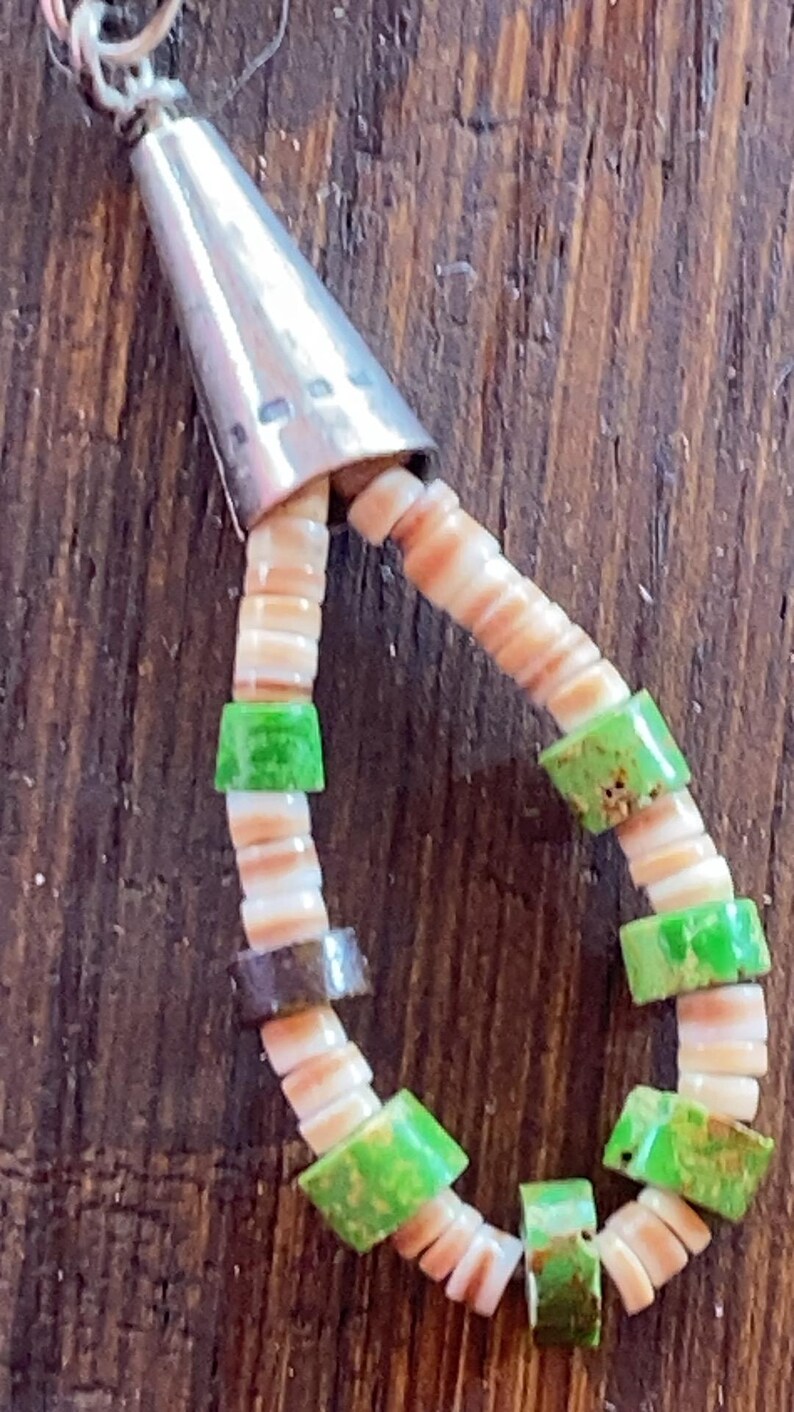 May include: A handcrafted necklace featuring a silver cone pendant and a beaded design. The necklace incorporates small, flat, off-white shell beads interspersed with vibrant green and brown square beads. The beads are strung together, creating a unique and eye-catching accessory.