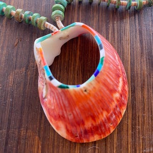 May include: A handcrafted necklace featuring a large, vibrant orange shell pendant with colorful accents. The shell is strung on a necklace of turquoise and tan-colored beads, creating a striking contrast. The necklace is displayed on a wooden surface.