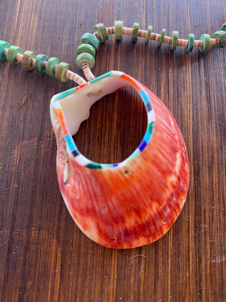 May include: A handcrafted necklace featuring a large, vibrant orange shell pendant with colorful accents. The shell is strung on a necklace of turquoise and tan-colored beads, creating a striking contrast. The necklace is displayed on a wooden surface.