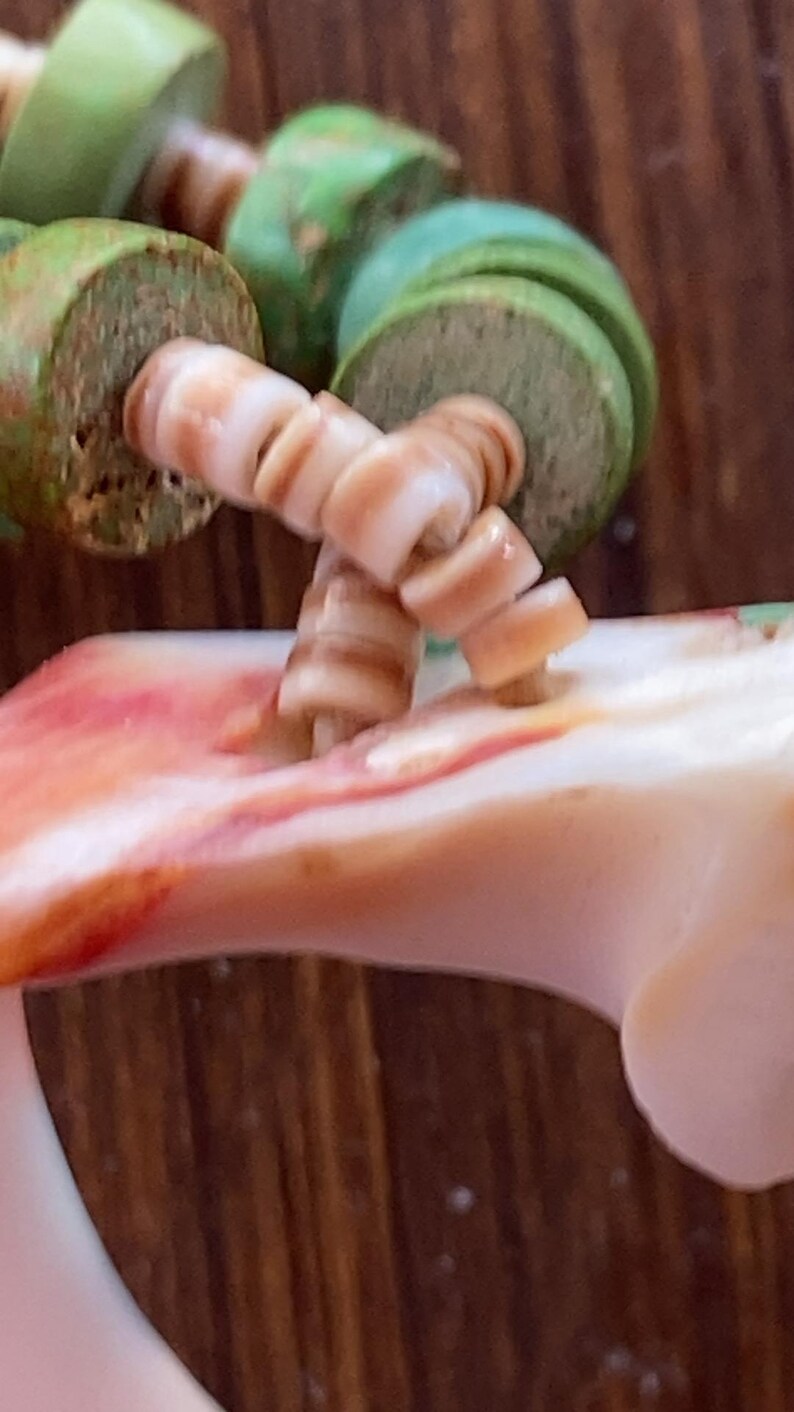 May include: Close-up of a handcrafted necklace featuring natural shell and green wooden beads. The shell beads are a light tan color, and the wooden beads are a muted green. The necklace is displayed against a blurred wooden background.