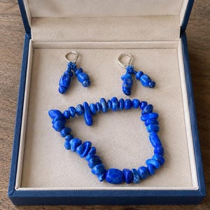 May include: A jewelry set featuring a bracelet and a pair of earrings, all made of vibrant blue stones. The set is presented in a blue box with a cream-colored interior, creating a striking contrast. The earrings have silver hooks.