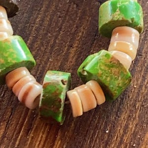 May include: Close-up of a beaded bracelet featuring alternating green and tan beads. The green beads are rectangular and have a distressed finish. The tan beads are shell-shaped and have a natural, organic look. The bracelet is on a wooden surface.