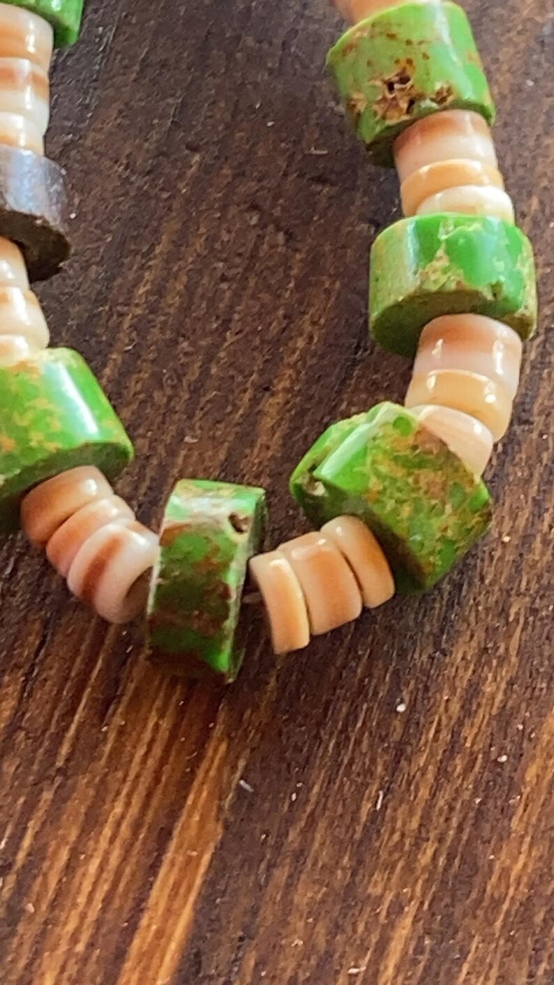 May include: Close-up of a beaded bracelet featuring alternating green and tan beads. The green beads are rectangular and have a distressed finish. The tan beads are shell-shaped and have a natural, organic look. The bracelet is on a wooden surface.