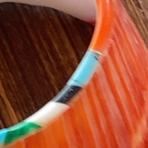 May include: Close-up of a colorful bracelet with a mix of colors. The bracelet features sections of orange, white, black, and turquoise. The design includes a striped pattern on the orange sections, creating a textured appearance. The bracelet is set against a wooden background.
