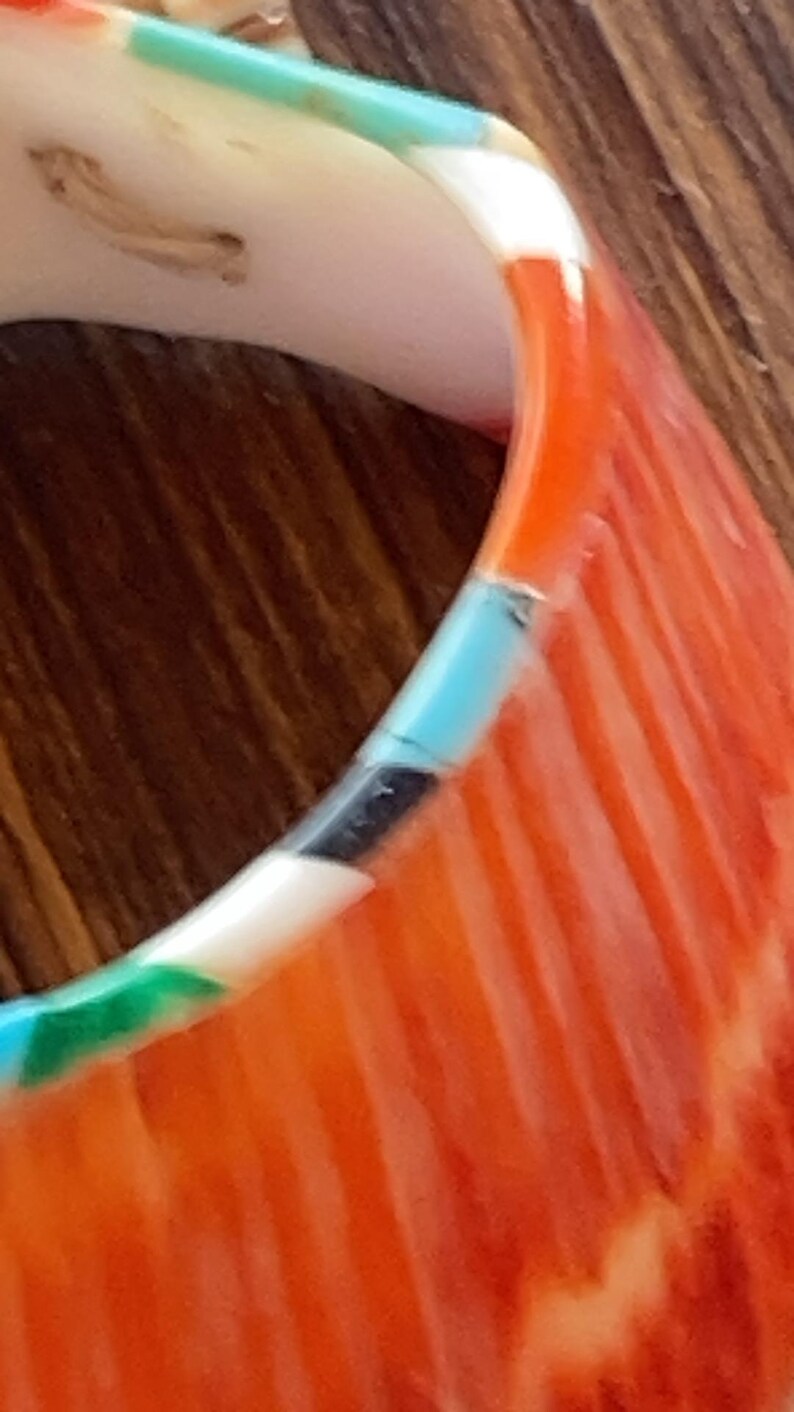 May include: Close-up of a colorful bracelet with a mix of colors. The bracelet features sections of orange, white, black, and turquoise. The design includes a striped pattern on the orange sections, creating a textured appearance. The bracelet is set against a wooden background.