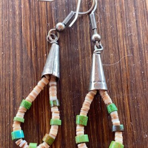 May include: Handmade earrings featuring a teardrop design. The earrings have silver-colored cone-shaped tops and are adorned with small, colorful beads in shades of green, tan, and turquoise. The earrings are on a wooden surface.