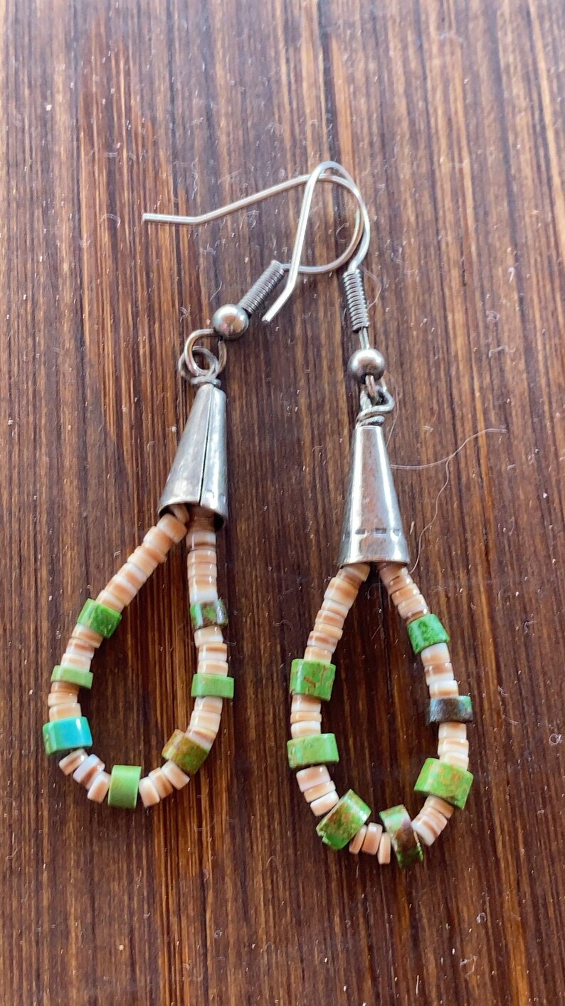 May include: Handmade earrings featuring a teardrop design. The earrings have silver-colored cone-shaped tops and are adorned with small, colorful beads in shades of green, tan, and turquoise. The earrings are on a wooden surface.