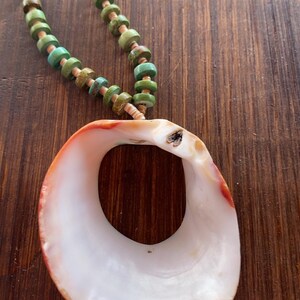 May include: A handcrafted necklace featuring a large, white seashell pendant with orange and red accents. The necklace has a beaded strand of turquoise and brown beads, creating a natural, bohemian aesthetic.