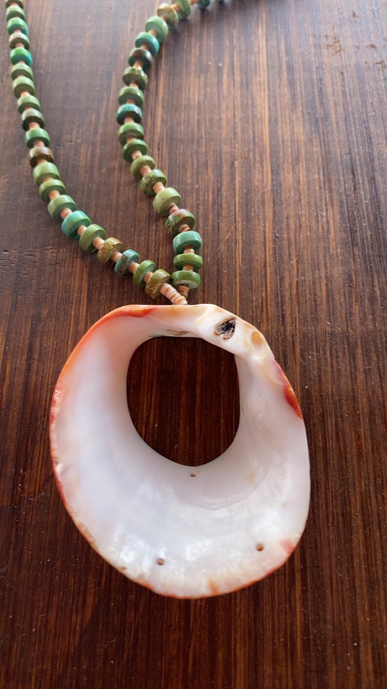 May include: A handcrafted necklace featuring a large, white seashell pendant with orange and red accents. The necklace has a beaded strand of turquoise and brown beads, creating a natural, bohemian aesthetic.