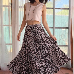 May include: A sleeveless, light pink crop top paired with a long, flowing skirt. The skirt features a black and white damask pattern. A long beaded necklace completes the outfit. The model is smiling.