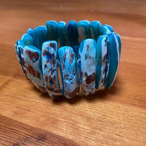 Op de afbeelding: Een blauwe en witte gepatroneerde armband gemaakt van meerdere schelpen. De armband is elastisch en kan worden uitgerekt om verschillende polsmaten te passen.